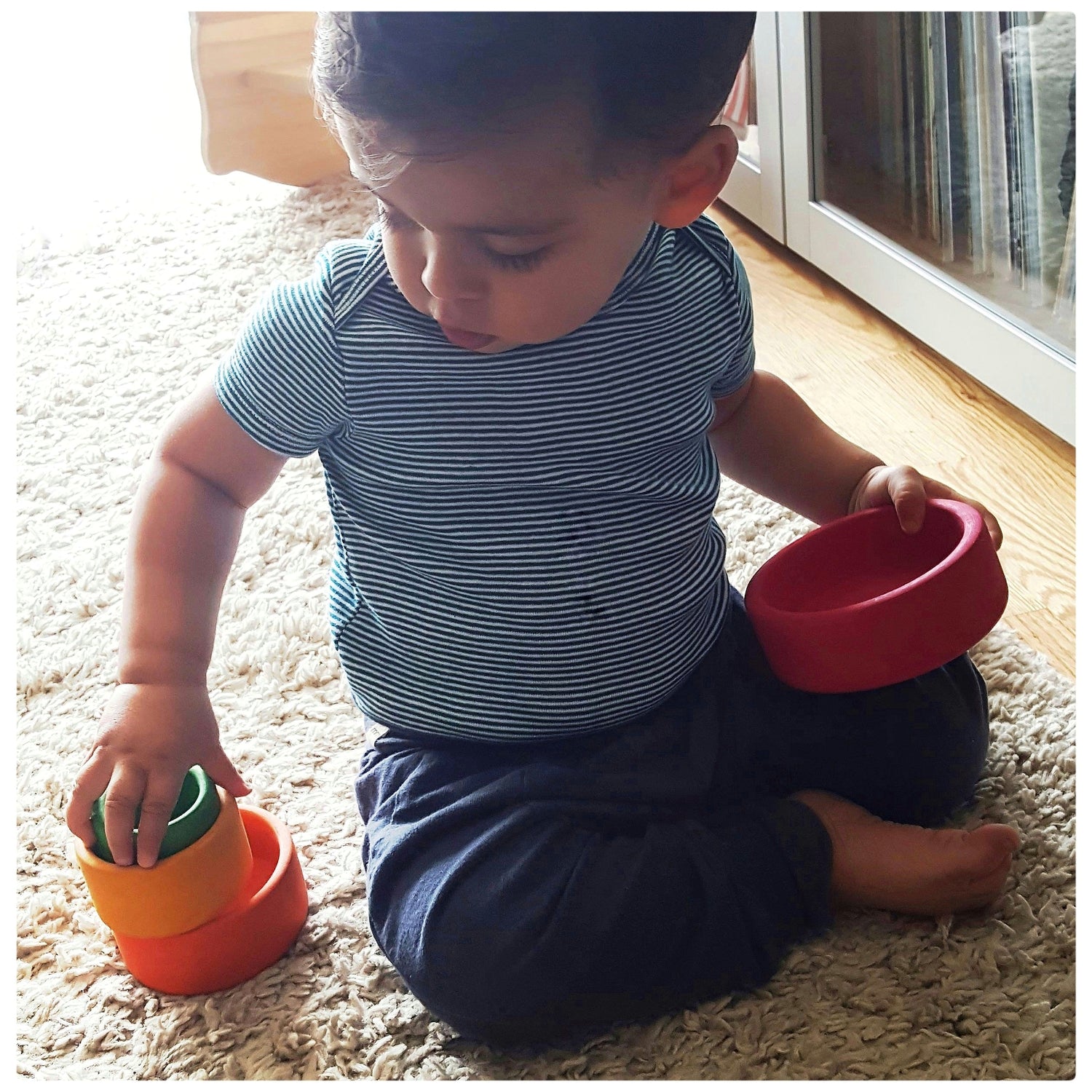 A 15 month old toddler sits on the floor and plays with a rainbow wooden nesting bowls toy from his toy subscription box.